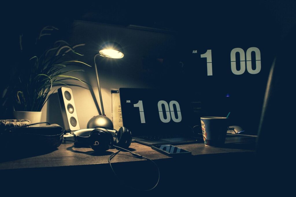A dimly lit modern office desk with a laptop, headphones, and speakers at 1:00 AM.